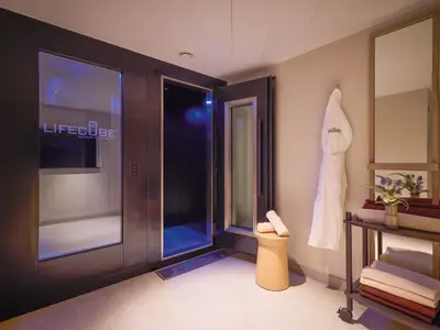 Blick in einen hellen Spa-Raum an Bord der Mein Schiff Flow mit einer geöffneten Kryotherapie-Kammer namens "LifeCube". Ein Bademantel und gestapelte Handtücher liegen auf einem Regal und einem Hocker bereit.