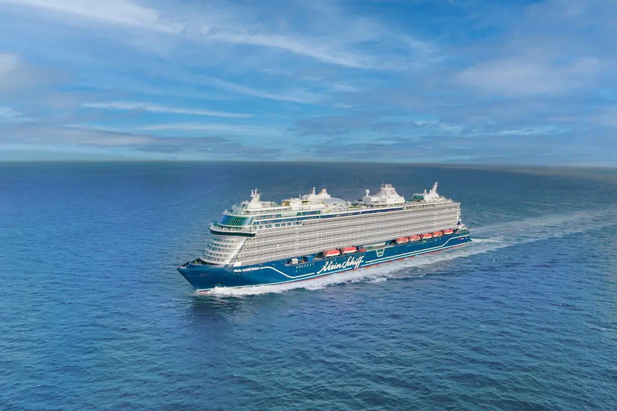 Luftaufnahme der Mein Schiff Relax auf dem offenen Meer. Das große, blau-weiße Kreuzfahrtschiff gleitet durch das Wasser und zieht eine Heckwelle. Blauer Himmel mit leichter Bewölkung.