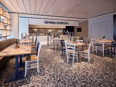 Innenansicht des Restaurants 'Taverna Dionysos' auf der Mein Schiff Relax. Helle Holztische mit blauen und weißen Stühlen stehen auf einem gemusterten Boden. Im Hintergrund ist eine offene Küche mit Edelstahlgeräten zu sehen, darüber leuchtet der Schriftzug 'Taverna Dionysos'.