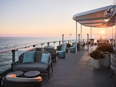 Ein luxuriöses Außendeck der Mein Schiff Relax bei Sonnenuntergang. Bequeme Outdoor-Möbel und Lounges laden zum Entspannen ein, mit Blick auf das ruhige Meer und den untergehenden Horizont.