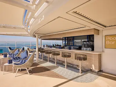 Blick auf die Pool Bar der Mein Schiff Relax, einer modernen Outdoor-Bar mit stilvollen Rattanhockern und blauen Details. Im Vordergrund sind bequeme Sessel mit blauen Kissen und kleinen Tischen zu sehen, dahinter erstreckt sich das weite, blaue Meer unter einem klaren Himmel.