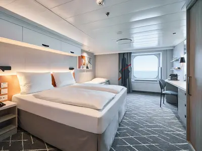 Moderne Außenkabine an Bord der Mein Schiff Relax. Doppelbett, Schlafsofa, Schreibtisch und Meerblick durch ein Bullaugenfenster. Helles Design mit orangefarbenen Akzenten.