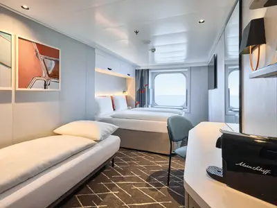 Helle und moderne Außenkabine an Bord der Mein Schiff Relax. Ausgestattet mit zwei Betten, Schreibtisch, Kaffeemaschine und großem Fenster mit Meerblick. Die Wände sind mit Kunstwerken verziert.