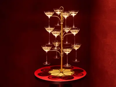 Ein goldener Sektkelchbaum mit zahlreichen gefüllten Champagnergläsern steht auf einem roten Sockel vor tiefroter Wand. Die festliche Präsentation ist an Bord der Mein Schiff Relax zu sehen.