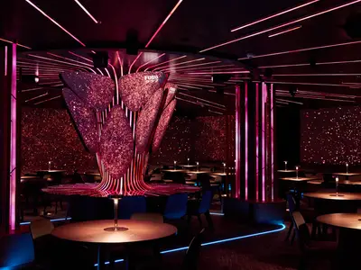 Die FUGU Bar & Lounge an Bord der Mein Schiff Relax. Pinke Neonlichter an Decke, Säulen und einer zentralen FUGU-Skulptur. Glitzernde Wandprojektionen und runde Tische mit Einzelbeleuchtung schaffen ein futuristisches Ambiente. Ohne Personen.
