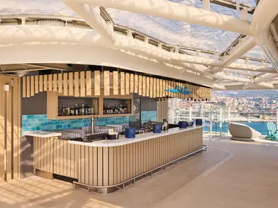 Eine moderne, überdachte Outdoor-Bar an Bord der Mein Schiff Relax mit Holzelementen und blauen Fliesen. Im Hintergrund sind eine Hafenstadt und das Meer bei Tageslicht zu sehen.