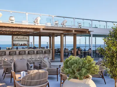 Die Agora Bar an Bord der Mein Schiff Relax ist ein stilvoller Außenbereich mit Barhockern, Loungebereichen, vielen Pflanzen und einem weiten Blick auf das offene Meer.