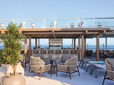 Die Agora Bar auf dem Sonnendeck der Mein Schiff Relax mit Bar, Barhockern, modernen Lounge-Sesseln und einem Pergola-Dach. Im Hintergrund das weite Meer.
