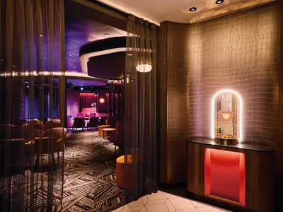 Eingang La Cage Lounge Mein Schiff Relax. Goldener Vogelkäfig mit Herz auf Sideboard vor strukturierter Wand. Vorhänge zur violett beleuchteten Bar mit Sitzgelegenheiten.