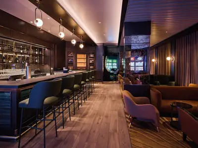 Elegante Bar-Lounge auf der Mein Schiff Relax. Eine dunkle Holztheke mit Hockern und gemütliche Sessel prägen den Raum, dahinter leuchten Casino-Spielautomaten.