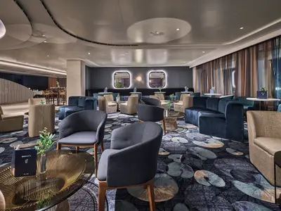 Ein moderner Loungebereich der Atlantik Bar an Bord der Mein Schiff Relax mit blauen und beigefarbenen Sitzgelegenheiten, Tischen und einem gemusterten Teppich. Im Hintergrund eine Bar und Fenster mit Vorhaengen.
