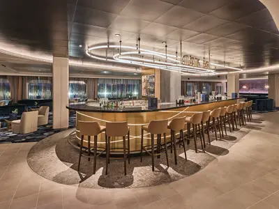 Die elegante Atlantik Bar an Bord der Mein Schiff Relax, gesehen aus dem Barbereich. Sie zeigt eine goldene Theke, Barhocker und gemütliche Sitzbereiche mit Fenstern im Hintergrund.
