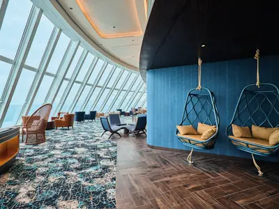 Heller Loungebereich an Bord der Mein Schiff Relax mit schrägen Panoramafenstern zum Meer. Blaue Hängesessel mit gelben Kissen stehen vor einer texturierten blauen Wand, davor Parkettboden. Links ein Bereich mit gemustertem Teppich und braunen Ledersesseln.