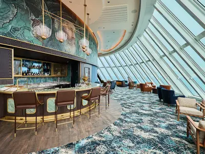 Eine moderne Bar-Lounge an Bord der Mein Schiff Relax. Die halbrunde Bar mit Marmor- und Holzverkleidung, goldenen Akzenten und Barhockern. Eine grosse Fensterfront bietet Meerblick. Die Wand ziert ein grosses blau-gruenes Wandgemälde.