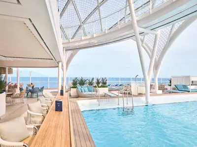 Blick auf den hellen, ueberdachten Aussenpoolbereich der Mein Schiff Relax mit blauem Meer im Hintergrund, Bar und Lounge-Sesseln.