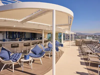 Moderne Poolbar mit blauen Akzenten und weissen Lounge-Sesseln unter einem Sonnendach auf dem Deck der Mein Schiff Relax. Hinten sind Liegen, ein Pool und eine Stadt mit Bergen sichtbar.