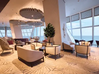Die moderne X-Lounge an Bord der Mein Schiff Relax. Große Fenster mit Meerblick. Helle Sitzgruppen, Tische, ein gemusterter Teppich und eine große Pflanze.