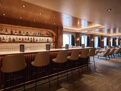 Elegante Bar-Lounge an Bord der Mein Schiff Relax. Lange Theke mit Barhockern und Spirituosenregalen. Dahinter Loungebereich mit Sesseln und Fenstern. Modernes Design mit Holz und Marmor.