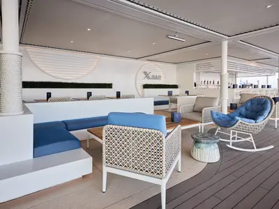 Die moderne X-Bar an Bord der Mein Schiff Relax, gefüllt mit blauen Sofas und Rattan-Sesseln. Die weiße Bar und der Meerblick im Hintergrund runden das Bild ab.