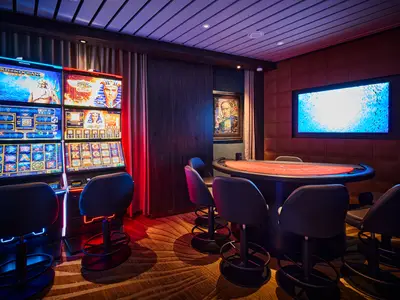 Das Casino der Mein Schiff Relax mit leuchtenden Spielautomaten und einem runden Pokertisch. Gedämpftes, farbiges Licht schafft eine spannende Atmosphäre. Wandkunst und ein Bildschirm sind sichtbar.