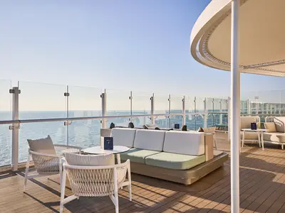 Sonnendeck der Mein Schiff Relax mit bequemen Loungemöbeln, darunter Sofas und Stühle in hellen Tönen, vor einer Glasbrüstung mit weitem Meerblick unter blauem Himmel. Eine überdachte Struktur bietet Schatten.