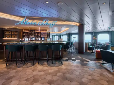 Eine moderne Bar an Bord der Mein Schiff Relax. Barhocker stehen vor der Theke mit "Mein Schiff"-Neonlicht. Große Fenster bieten Meerblick, dahinter bequeme Sessel.