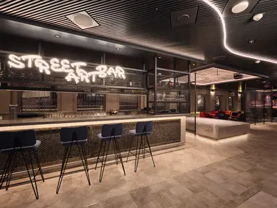 Blick in die moderne Street Art Bar an Bord der Mein Schiff Relax. Ein leuchtender Neon-Schriftzug 'STREET ART BAR' hängt über der Theke. Im Vordergrund sind vier blaue Barhocker zu sehen, dahinter eine lange Bar mit Gläsern. Im Hintergrund befinden sich gemütliche Sitzgelegenheiten in Rot und Schwarz und eine Wand mit Graffiti-Kunst.