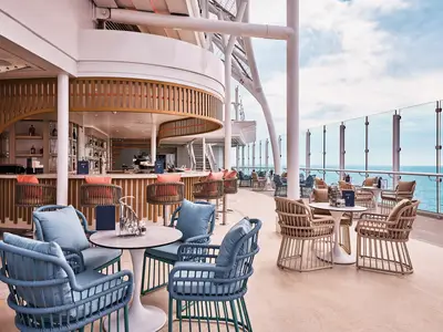 Blick auf die stylische Außenbar und Sitzbereiche auf dem Deck der Mein Schiff Relax, mit Meerblick durch eine Glasbrüstung. Helle Möbel und Holzverkleidung schaffen eine entspannte Atmosphäre.