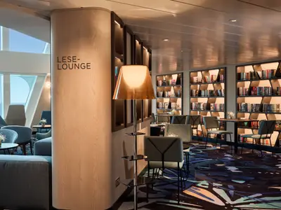 Innenansicht der Leselounge an Bord der Mein Schiff Relax. Helle Räumlichkeiten mit komfortablen Sitzecken, grossen Panoramafenstern mit Meerblick und eleganten, beleuchteten Bücherregalen mit vielen Büchern.