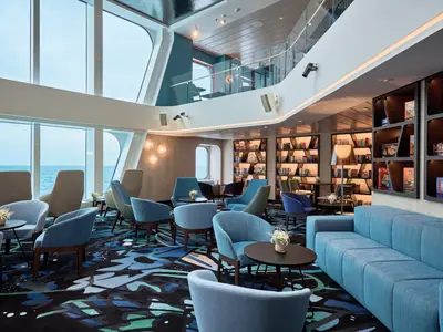 Die helle Leselounge an Bord der Mein Schiff Relax mit grossen Fenstern zum Meer. Blaue Sessel und Sofas umgeben Buecherregale und Brettspiele.