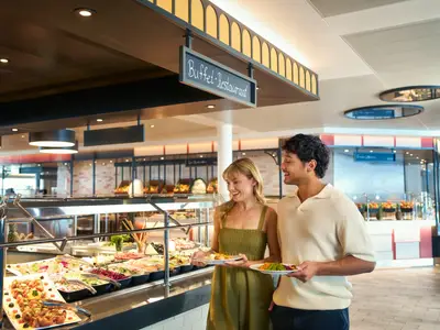 Ein laechelndes Paar mit Tellern waehlt Speisen von einem reichhaltigen Buffet im Buffet-Restaurant der Mein Schiff Relax. Verschiedene Salate und warme Speisen sind ausgestellt.