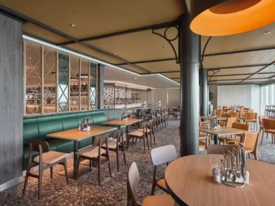 Innenansicht des Harbour Market Buffet-Restaurants auf der Mein Schiff Relax mit zahlreichen Holztischen, Stühlen und einer grünen Sitzbank. Im Hintergrund ist die Buffettheke zu sehen.