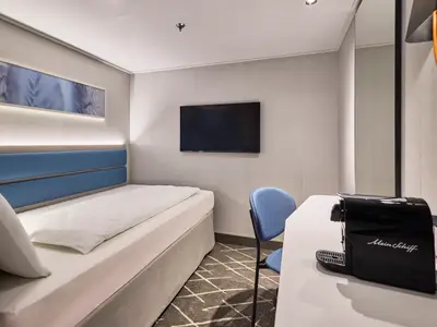 Blick in eine moderne Einzelkabine auf der Mein Schiff Relax. Ein einzelnes Bett mit blauem Kopfteil, ein Flachbildfernseher und eine Kaffeemaschine auf einem Schreibtisch sind zu sehen.