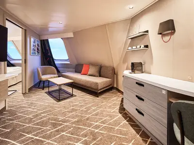 Ein hell eingerichteter Wohnbereich in einer Familienkabine an Bord der Mein Schiff Relax ist zu sehen. Im Vordergrund ein Sofa mit Kissen, ein Couchtisch und ein Sessel. Durch die ovalen Fenster ist blauer Himmel und Meer sichtbar. Eine Kaffeemaschine steht auf einer Kommode.