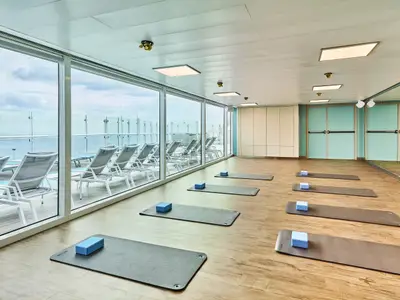Ein heller Kursraum an Bord der Mein Schiff Relax mit großen Fenstern zum Meer. Yoga-Matten und Blöcke liegen auf dem Holzboden bereit. Draußen sind Liegestühle auf einem Deck sichtbar.
