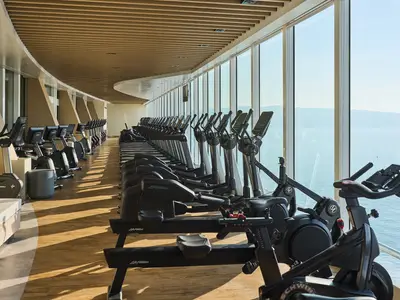 Ein heller, langer Fitnessraum an Bord der Mein Schiff Relax. Reihen von Laufbändern, Crosstrainern und Fahrrädern stehen vor großen Fenstern mit Meerblick.