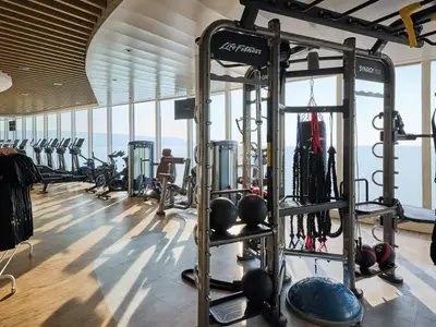 Heller Fitnessbereich an Bord der Mein Schiff Relax mit modernen Kraft- und Kardiogeraeten. Durch grosse Fenster bietet sich ein weiter Blick auf das Meer und die Kueste.