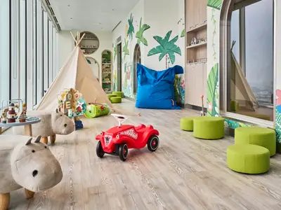 Ein farbenfroher Babyraum an Bord der Mein Schiff Relax mit einem Spielzelt, Plüsch-Hippos und grünen Hockern, umgeben von Dschungel-Wandmalereien und großen Fenstern.