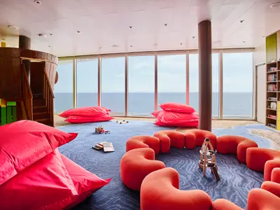 Bunter Kinderbereich "Insel der Seeräuber" an Bord der Mein Schiff Relax mit Holzschiff, roten Sitzsäcken, orange Sitzmöbeln und Büchern. Große Fenster zum Meer.