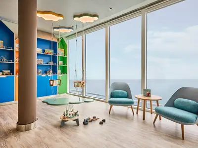 Der Kinderbereich 'Nest' an Bord der Mein Schiff Relax zeigt bunte Regale voller Spielzeug, zwei Babyschaukeln und helle Sessel mit Meerblick durch große Fenster.
