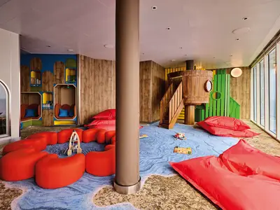 Der Spielbereich 'Insel der Seeräuber' an Bord der Mein Schiff Relax mit großer Holzhütte, Treppe, gemütlichen Sitzkojen und roten Sitzkissen. Ein blauer Teppich mit Wellenmuster und große Fenster mit weitem Meerblick füllen den Raum.