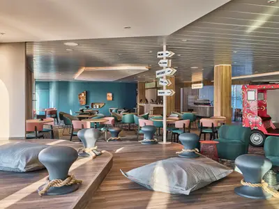 Ein heller, moderner Lounge-Bereich an Bord der Mein Schiff Relax mit verschiedenen Sitzgruppen, einer Destinationstafel und einem roten Tuk-Tuk im Hintergrund. Tageslicht erhellt den Raum.