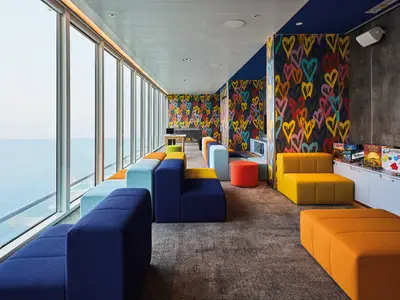 Lounge auf Mein Schiff Relax. Bunte, modulare Sitzmoebel in Blau, Gelb, Orange. Grosse Fenster zum Meer. Wand mit Herz-Graffitis. Regal mit Brettspielen.