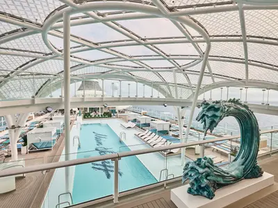 Der moderne Aussenpoolbereich der Mein Schiff Relax. Liegeflächen, Cabanas und eine Wellenskulptur unter Glasdach-Architektur. Weites Meer im Hintergrund.