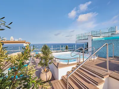 Helles Sonnendeck der Mein Schiff Relax mit einem sprudelnden Whirlpool, Stufen und Geländern aus Edelstahl. Hinten ein blauer Pool und das offene Meer unter blauem Himmel.