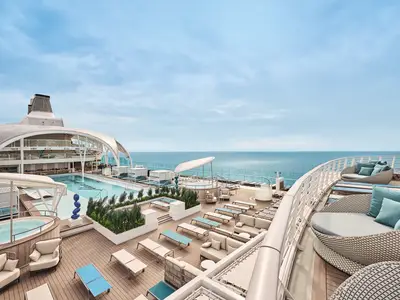 Blick auf den grossen Aussenpool und Whirlpools auf dem Sonnendeck der Mein Schiff Relax. Zahlreiche Liegen und Loungemoebel laden zum Entspannen ein. Im Hintergrund das weite Meer.