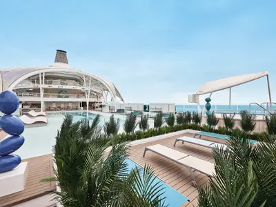 Blick auf den grosszuegigen Aussenpoolbereich an Bord der Mein Schiff Relax. Moderne Architektur, blaue Skulpturen und viele Pflanzen umgeben den Pool mit Liegen.