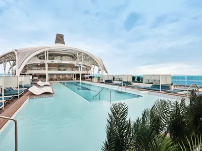 Aussenpool an Bord der Mein Schiff Relax. Moderne Liegen, Cabanas und eine geschwungene Glas-Architektur umgeben den langen Pool. Im Hintergrund das Meer.
