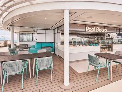 Das Pool Bistro auf dem Aussendeck der Mein Schiff Relax. Türkisfarbene Stühle und Tische auf Holzdeck, daneben Buffetbereich. Im Hintergrund das offene Meer.