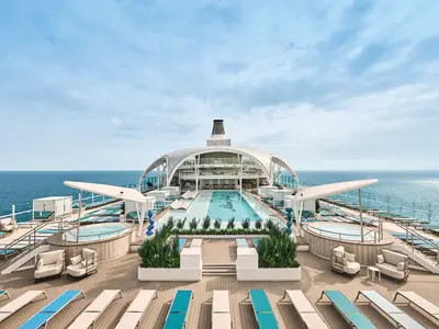 Blick auf das grosszügige Pooldeck der Mein Schiff Relax. Ein langer Pool, umgeben von Liegestühlen und Whirlpools. Im Hintergrund das offene Meer unter blauem Himmel.